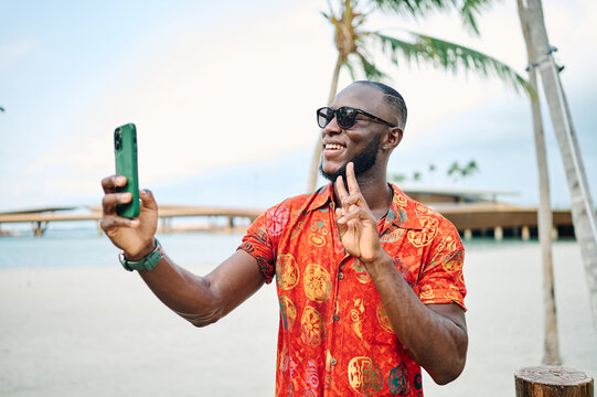 A smiling man takes a selfie on a tropical beach lined with palm trees while wearing sunglasses and a bright patterned shirt. He holds a smartphone, enjoying a relaxed vacation moment at a sunny resor
