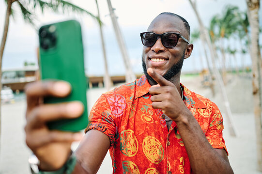 A smiling man takes a selfie on a tropical beach lined with palm trees while wearing sunglasses and a bright patterned shirt. He holds a smartphone, enjoying a relaxed vacation moment at a sunny resor