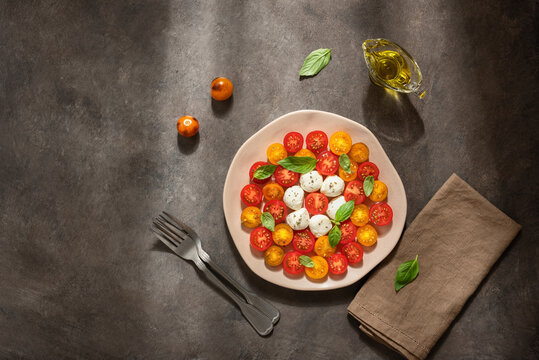 Caprese salad with mozzarella, colorful cherry tomatoes and basil leaves in a plate on a dark rustic background in sunlight, top view, flat lay