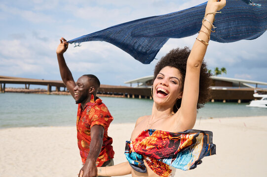 A joyful couple runs hand in hand along a sandy beach near a yacht and modern marina, laughing and waving a blue scarf overhead. Warm tropical palms, bright clothing and carefree movement convey roman