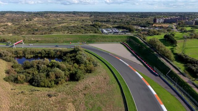 High-angle aerial drone view of Circuit Zandvoort race track wide sweeping turn with sports cars driving near gravel runoff tire barriers and coastal dunes in North Holland Netherlands