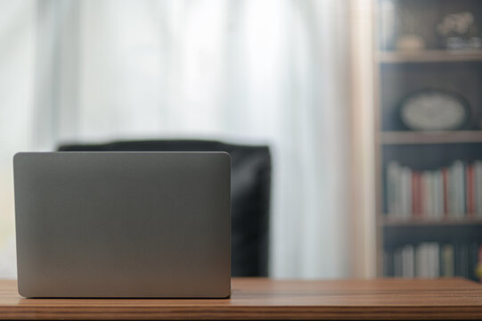Modern Closed Laptop on Wooden Desk in Bright Room with Bookshelf and Curtains