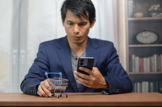 Young man using smartphone with miniature shopping cart on wooden table in modern home
