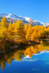 Fototapeta premium Vertical photo. Mountain landscape with a calm lake reflecting autumn trees with colorful fall foliage. Clear daytime view of water reflection, forest, and mountain peaks.