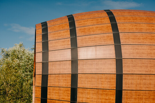 close-up of curved wooden exterior cladding with vertical black metal straps against blue sky and adjacent tre