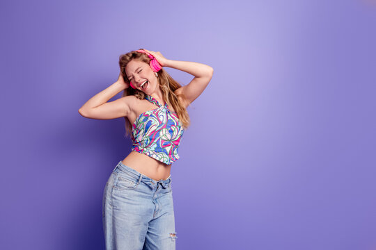 Young woman in colorful top and jeans dancing and enjoying music with pink headphones against a purple background