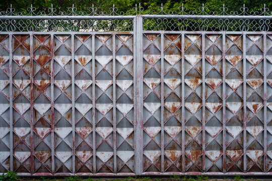 Rusty metal gate with geometric diamond pattern and decorative top railing, showing weathered texture and aged appearance in outdoor setting