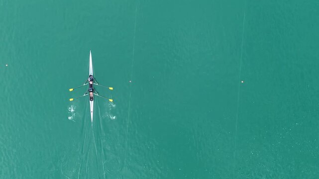 Top-down drone footage of a double sculls crew rowing across the turquoise waters of Lake Bled, Slovenia, with oars splashing and leaving ripples on the water.