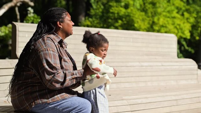 African American mother relaxing on park bench and talking with toddler daughter. Footage suitable for parenting campaigns, family lifestyle ads, and maternal wellness content
