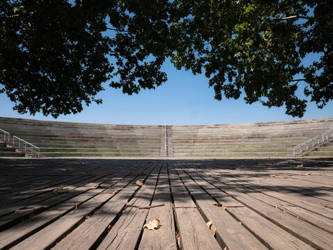 View of the open-air amphitheater in Madrid's Manzanares Linear Park, featuring curved tiered seating with wooden benches, staircases with railings, and a spacious stage with a wooden floor
