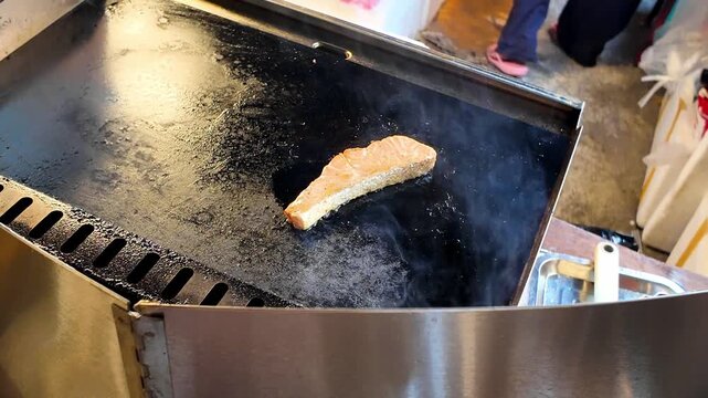 Close up shot of a flipped salmon fish steak with golden crispy crust frying on a hot flat iron griddle at a street food market stall in Thailand.
