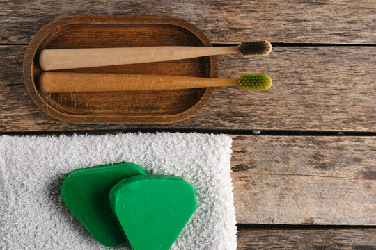 Flat lay of wooden toothbrushes in dish, green solid shampoo, and towel on wood.