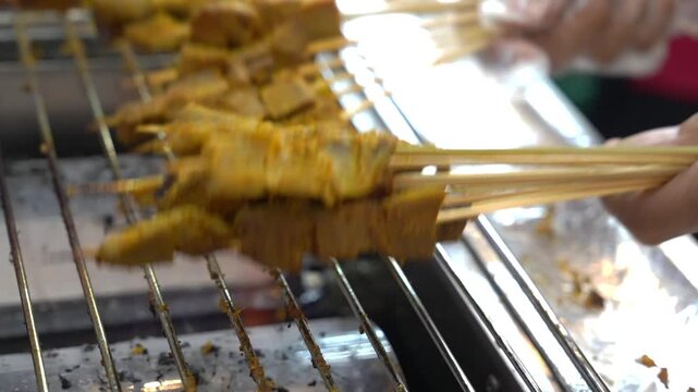 hand holding beef padang satay skewers with yellow curry marinade cooking on grill rack at street food stall.