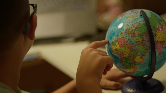 Young boy examines globe with curiosity. He wears glasses and touches continents thoughtfully. Globe spins slowly as he explores geography. Warm indoor light highlights his focused expression