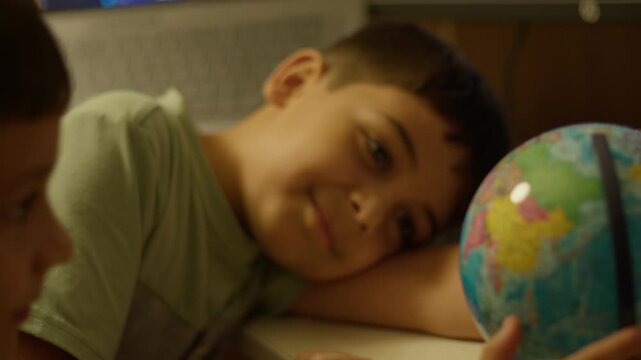 Curious child holds globe with wonder. Another child watches nearby, smiling. Warm indoor light, desk, laptop, globe. Ideal for education, travel, childhood learning themes