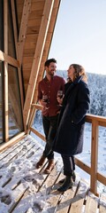 Fototapeta premium Casual winter balcony portrait of smiling couple with wine glasses at a mountain lodge for romantic getaway background and travel design