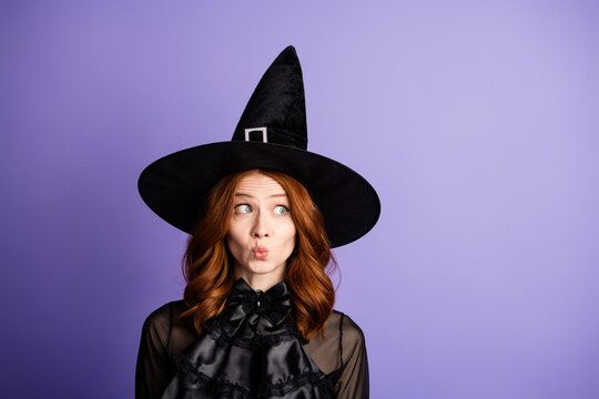 Playful redhead woman dressed as a witch in a black hat making a puckered funny face against a purple studio background for Halloween costume portrait and whimsical seasonal mood