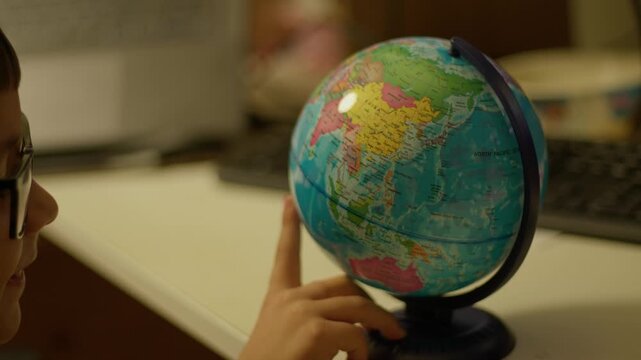 Globe held by child's hands. Child wearing glasses, studying continents. Soft light illuminates globe's surface. Child appears curious and engaged. Perfect for educational or travel-related projects