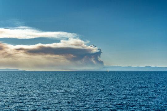 Large wildfire smoke plume rising over the sea with a distant sailboat on the horizon