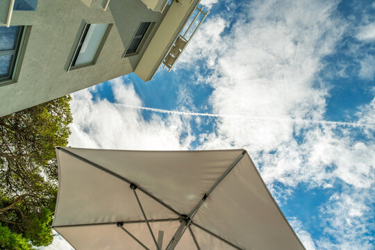 Low angle view of modern building facade, white sun umbrella, pine tree, and blue sky with white clouds and airplane contrail