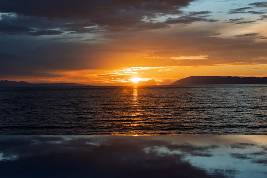 Stunning sunset over the sea with calm water, mountains in the distance, and dramatic clouds in the sky