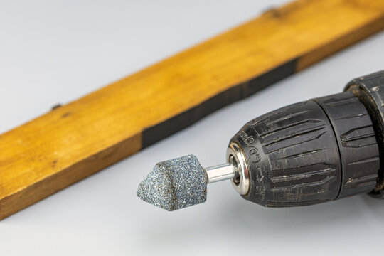 Professional macro photograph of a black cordless power drill keyless chuck holding a conical blue abrasive grinding stone bit for sharpening and polishing, with a blurred wooden tool handle
