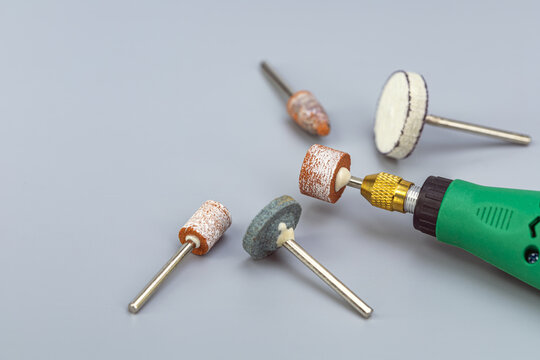 Close-up macro view of green electric mini rotary tool with golden brass collet chuck surrounded by assorted abrasive sanding drum bits, grinding stone wheels and white felt polishing attachment