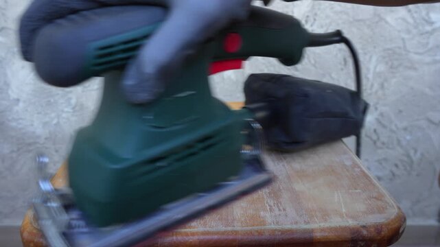 Close-up of an electric orbital sander smoothing the worn surface of an old wooden stool. The sanding process removes old finish and uneven patches, preparing the wood for restoration