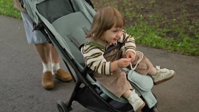 Caucasian toddler sitting in stroller holding beads while Caucasian mother pushes carriage in park, camera tilting up. Footage fits baby product advertising and parenting lifestyle campaigns