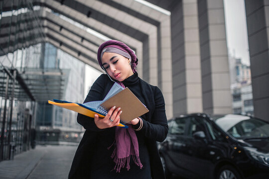 Busy young arabian woman stand outside. She hold yellow folde with documents and look down. Model talks on phone. She stand close to black cars.