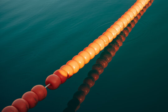 Diagonal line of orange plastic safety buoys floating on calm turquoise sea water with clear reflections.
