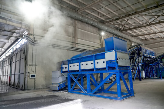 Large blue waste treatment machinery operates inside a high industrial hall as white vapor rises near ventilation ducts, illustrating equipment used in modern waste processing.