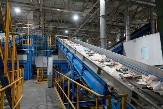 A blue conveyor belt carries mixed rubbish through an indoor sorting facility, with metal platforms, safety railings and overhead lights supporting industrial recycling operations.