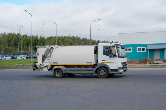A white municipal garbage truck drives along a paved road beside an industrial building, representing city sanitation services, refuse collection and everyday waste transport.