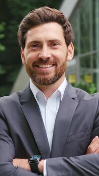Confident adult businessman in a formal suit standing outdoors near a modern glass office building and green park area. Smiling professional man looking directly at the camera with crossed arms