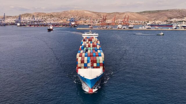Aerial drone cinematic video of truck size container cargo vessel leaving loading - unloading logistics port of Perama