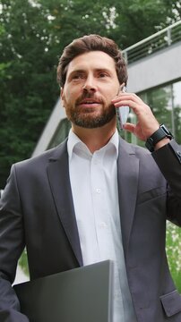 Successful businessman talking on phone while walking with laptop outdoors