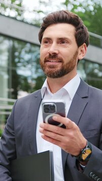Professional businessman standing outdoors near a modern office building, using a smartphone while holding a digital tablet. Thoughtful and focused male executive looking away as if considering
