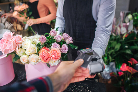 Cur view of man holding credit card in hand. He give it to florist. Flower seller has thereminal for cards. Table ull of colorful flowers.