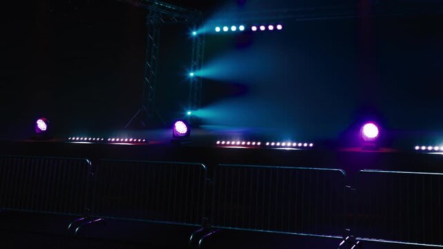 A beautifully lit stage awaits an energetic performance. Colorful beams of light shine down, creating an electrifying atmosphere, while barriers line the front, ready for eager fans.