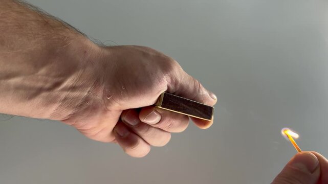 Hand holding a matchbox, lighting a match, flame ignition, close‑up of hands, dramatic lighting, minimalist background, moment of ignition, manual fire starting, tactile interaction.