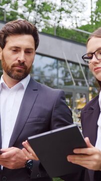 Businessman and businesswoman walk to the office while having a business conversation and looking at a tablet held by the woman, with the man carrying a laptop. Slow motion