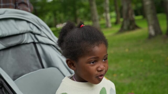 African American child sitting in stroller while young adult mother walks in park, camera tilting up. Footage suitable for parenting campaigns and family lifestyle advertising