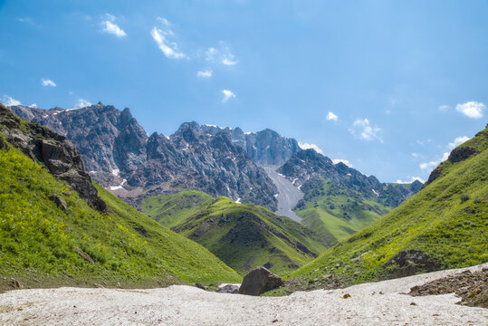 Vast alpine valley framed by green slopes and jagged rocky peaks, with lingering snow patches in the mountains near Badak Lake, Uzbekistan.