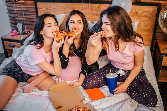 Three hungry teenagers biting pieces from pizza. They devour it. Hungry models wear pajamas. They sit on bed in room.