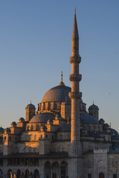 New Mosque (Yeni Cami), a 1665 Ottoman landmark in Istanbul, Turkey, its minaret and cascading domes glowing in golden hour light against a clear blue sky