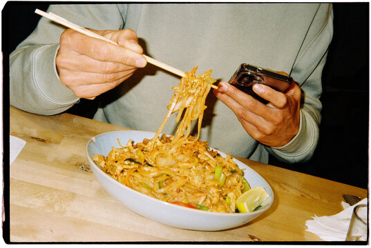 a man eats pad thai with chopsticks while holding a smartphone,