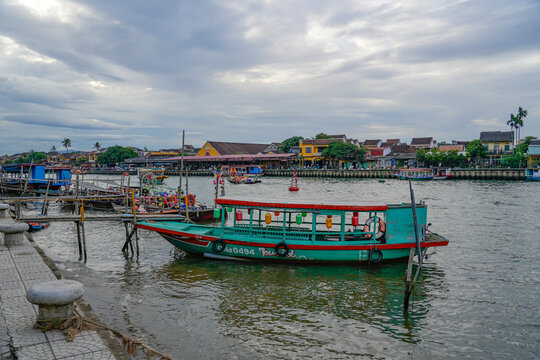 A traditional green wooden passenger boat moored on the Thu Bon River with Hoi An old town houses and waterfront activity behind it, Vietnam, Hoi An, July 7, 2026.