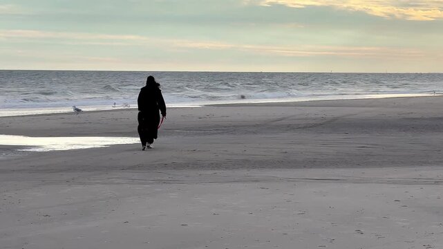 Girl in a long coat walking her dog along the ocean on a deserted beach on a windy winter day