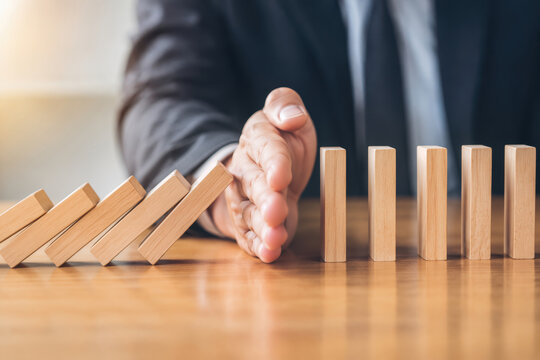 Businessman hand stopping falling wooden dominoes on table to prevent chain reaction corporate crisis prevention and strategic risk management concept for continuity and stability planning